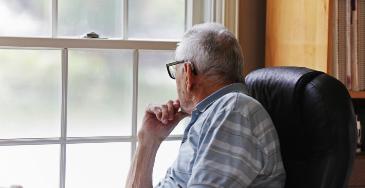 An older man is sitting quietly with his hand on his chin, looking out the window.