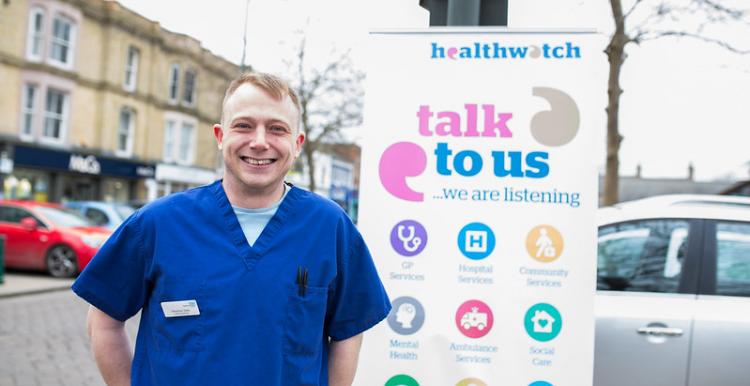 doctor stood in front of a talk to us us banner from Healthwatch