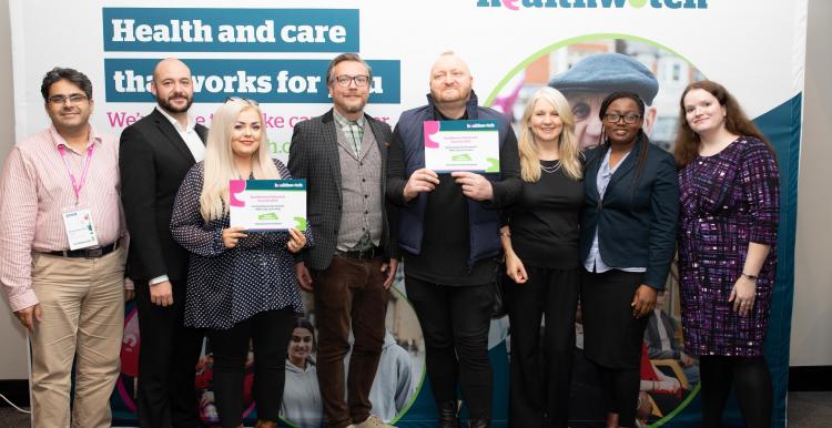 Group of adults stood smiling at the camera in front of a Healthwatch board