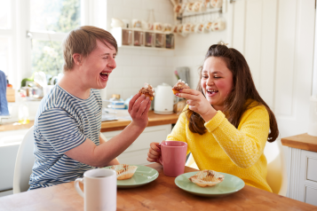 A young man with a learning disability enjoying tea and cake with a woman over a kitchen table.