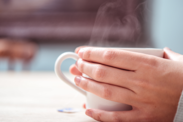 A person's hands wrapped around a mug. Steam is rising from the hot drink.