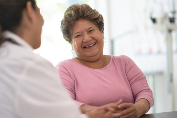 A woman speaking to a doctor. She is smiling and wearing a pink jumper.