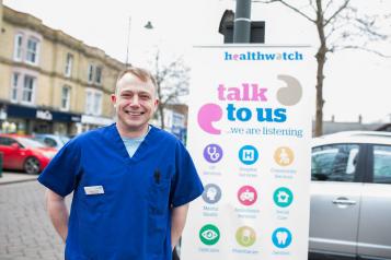 doctor stood in front of a talk to us us banner from Healthwatch