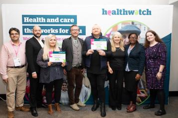 Group of adults stood smiling at the camera in front of a Healthwatch board