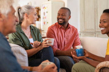 Four adults sitting in a living room, smiling, chatting, and drinking from mugs
