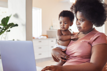 A Black mother holding her baby and looking at a laptop