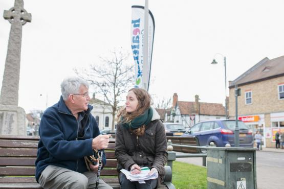 A man and woman sit talking on a bench in front of a vertical Healthwatch banner