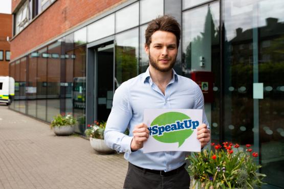 A young white man standing outside a hospital holding a sign which reads 'speak up'