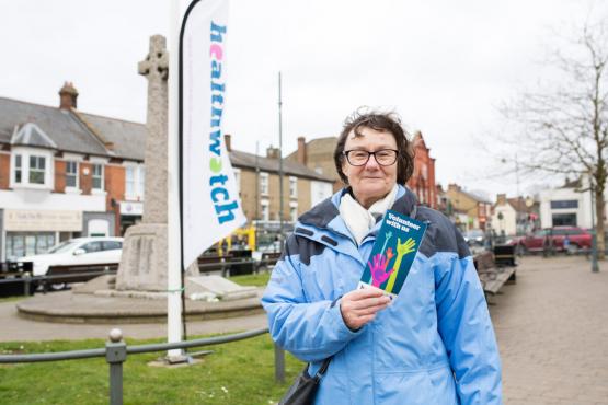 a woman standing in front of a Healthwatch banner, holding a Healthwatch leaflet