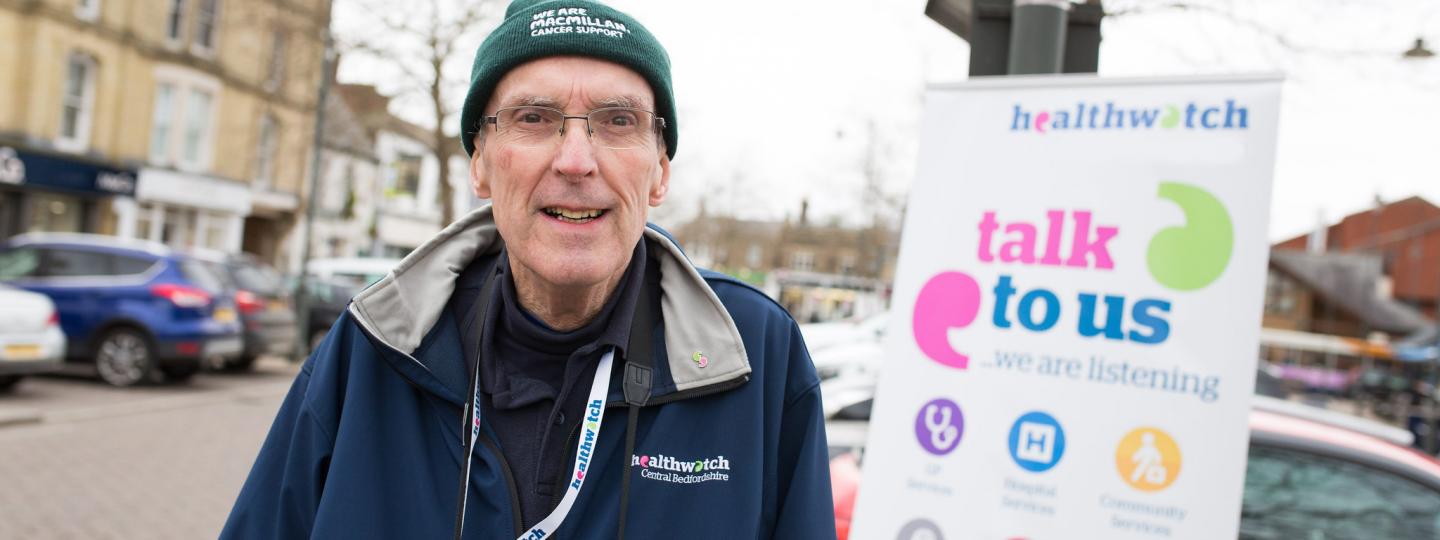 Man standing infront of a Healthwatch branded banner that says 'talk to us'