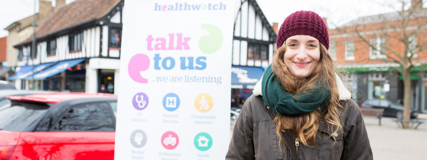 A woman wearing a hat and scarf, standing outside in front of a vertical Healthwatch banner