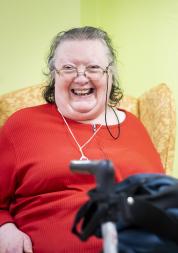 An older woman sitting in an arm chair and smiling. In front of her is a walking frame.