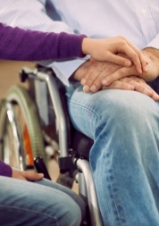 A woman sitting with her hand on top of a man's hands. The man is in a wheelchair.